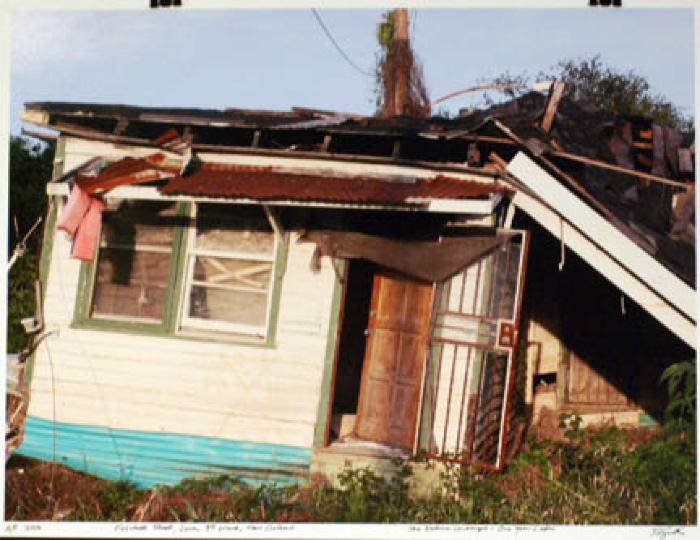 From the series of photographs by Steven L. Smith; image of a destroyed house with green trim around the doors and windows. Steel on the overhang.�� Severe roof damage and foundation damage based no the tilt of the home.